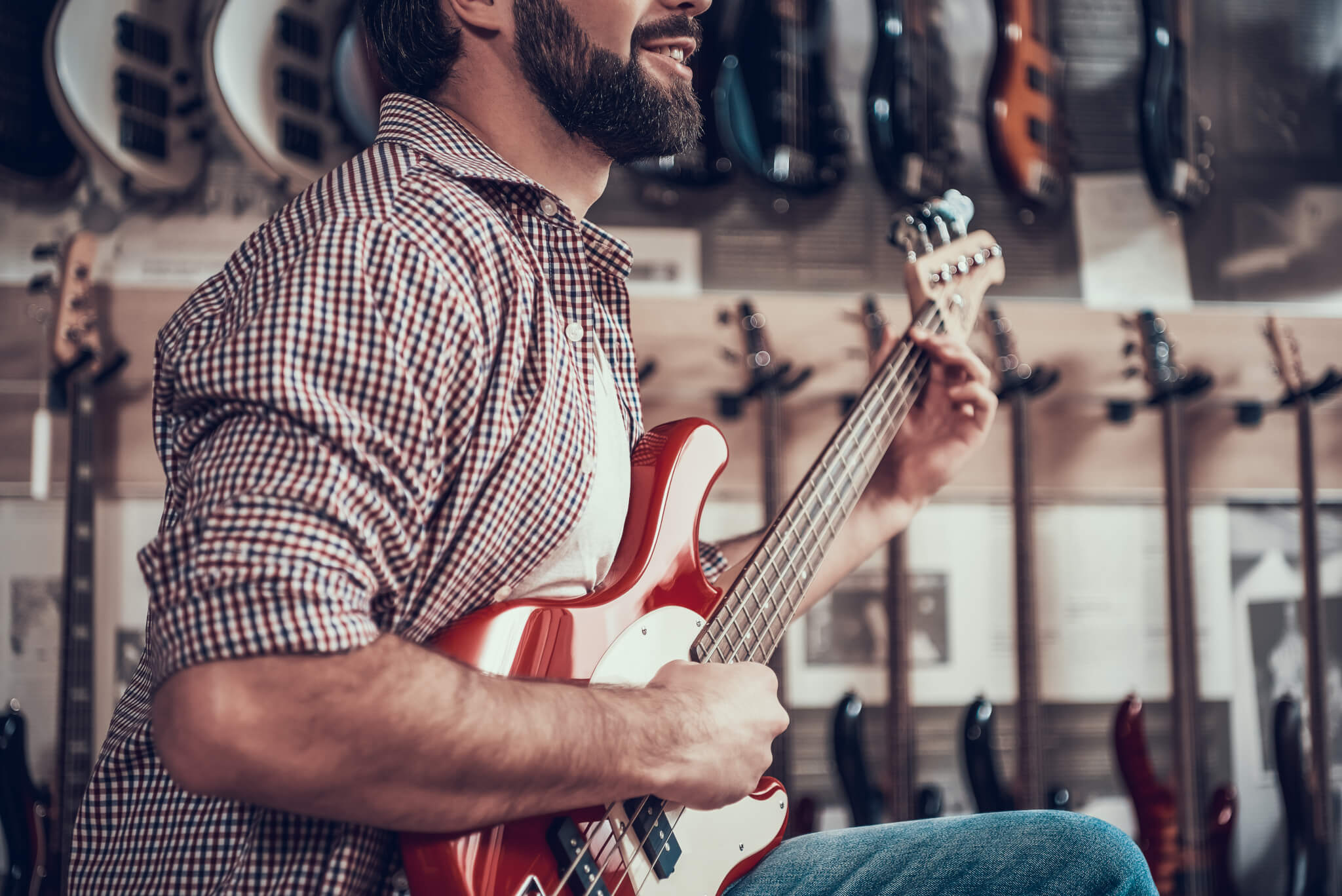 Bearded adult man plays on red electric guitar in musical instrument store.